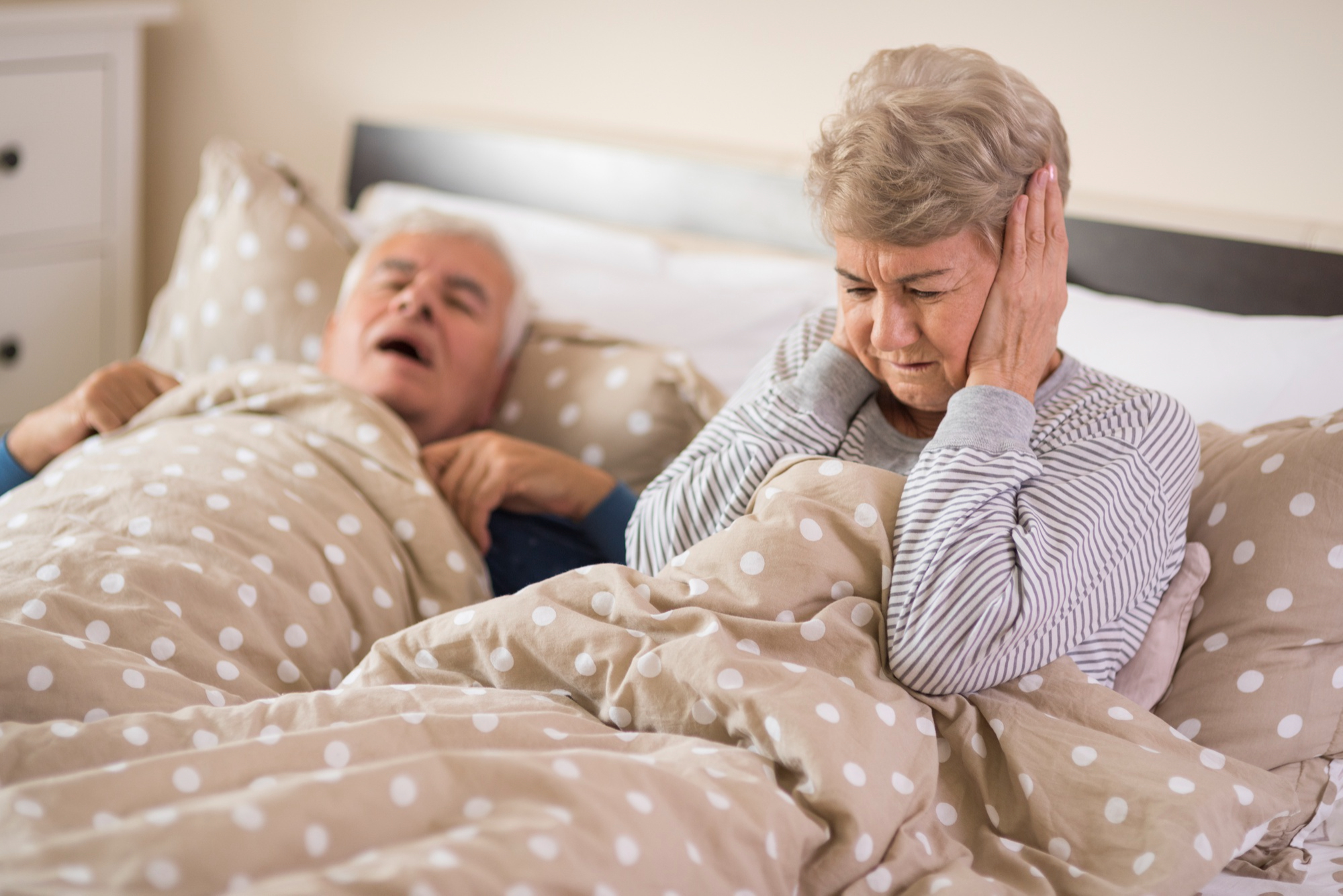 Senhora coloca as mãos no ouvido em sinal claro de incômodo por conta do ronco do companheiro, que tem apneia obstrutiva do sono.