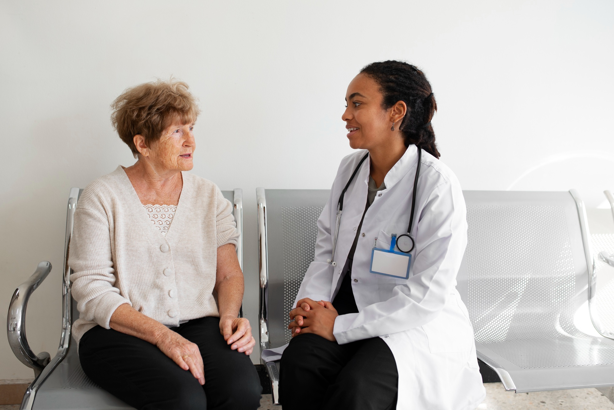 médica de família conversa com paciente idosa em sala de espera, demonstrando acolhimento e vínculo no atendimento.