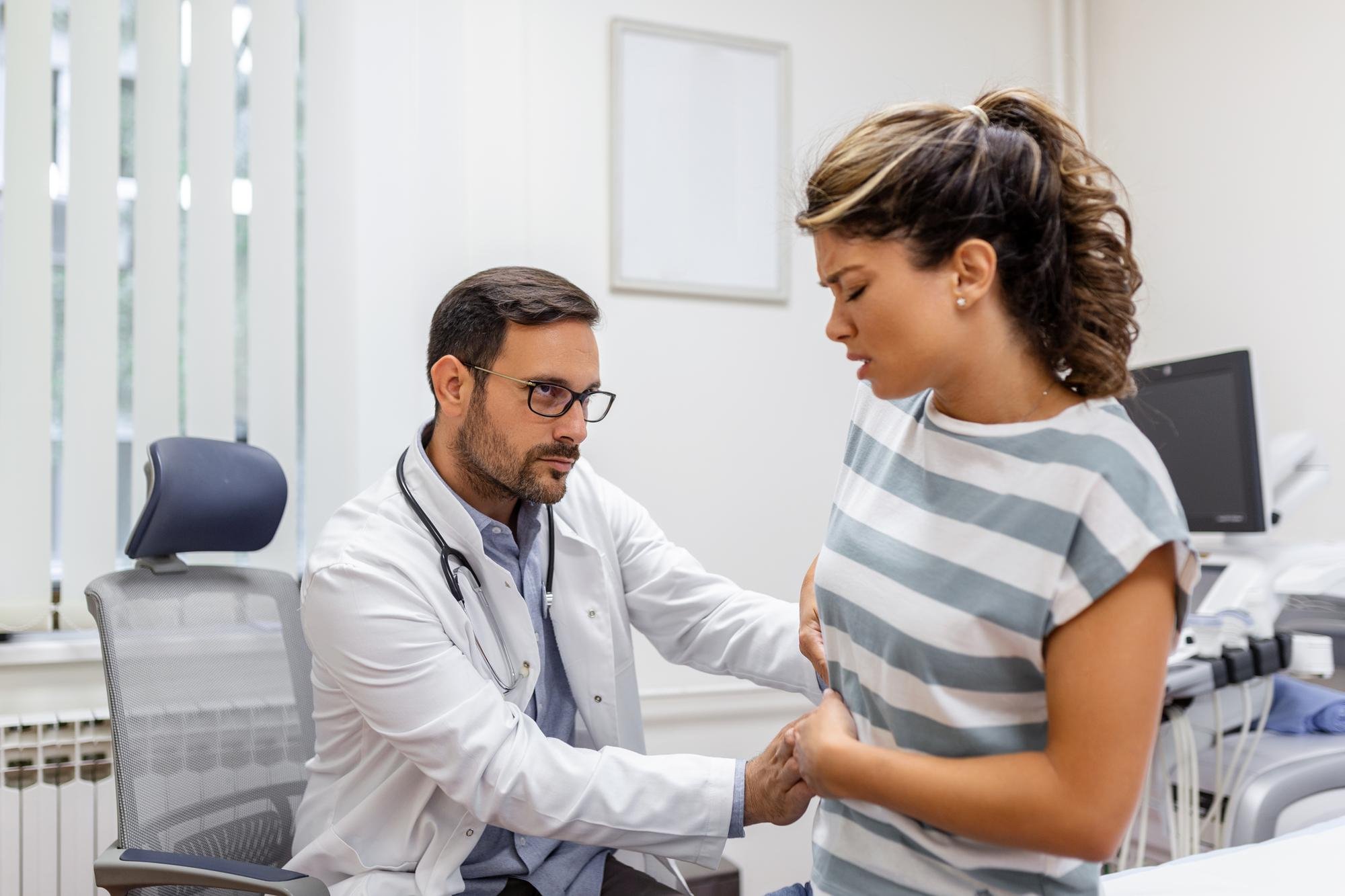Médico examina mulher jovem com dor abdominal e suspeita de doença da vesícula biliar.