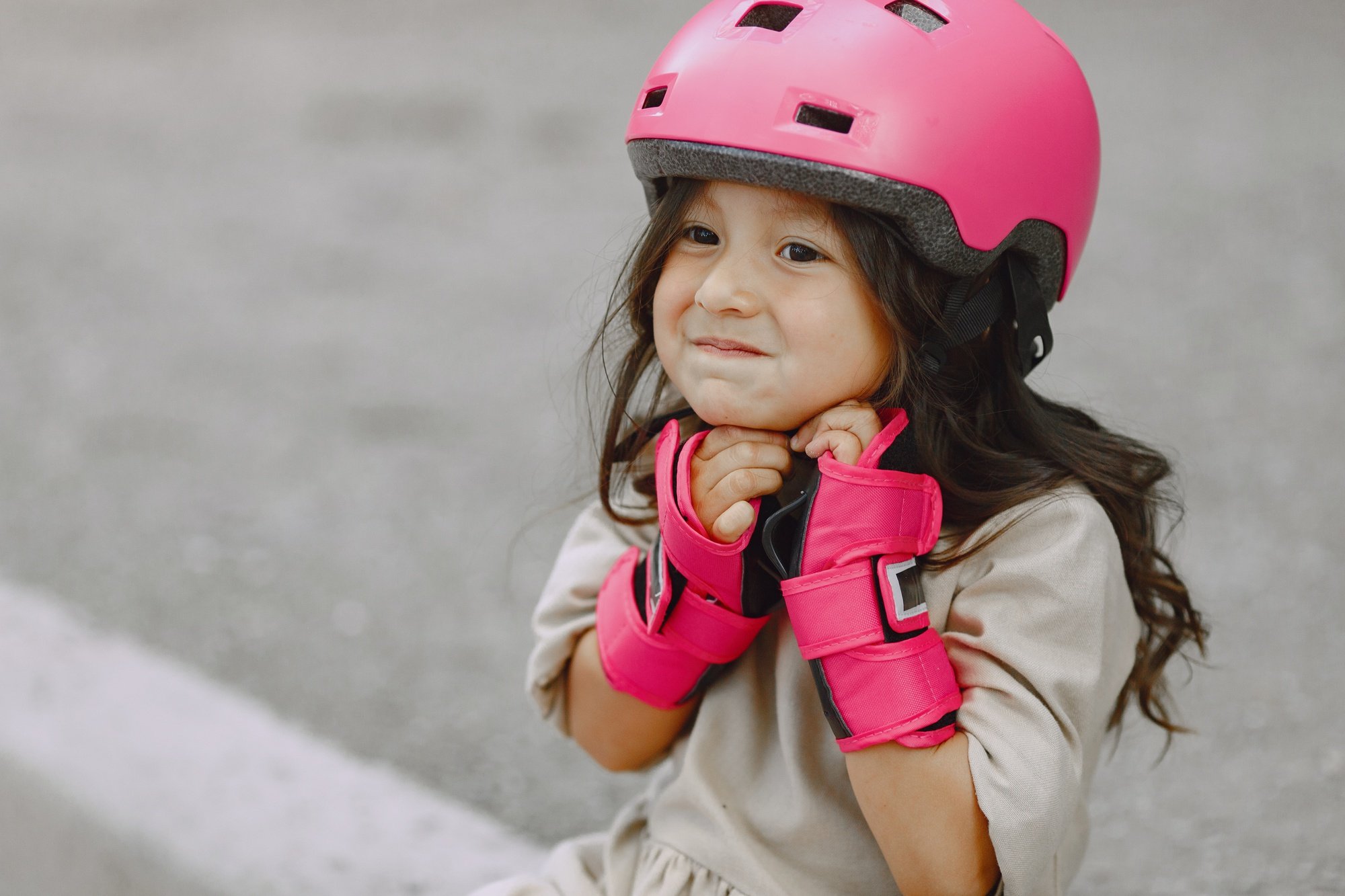 Menina sorri enquanto ajusta o capacete para criança rosa, usando luvas de proteção combinando, pronta para uma atividade ao ar livre com segurança.