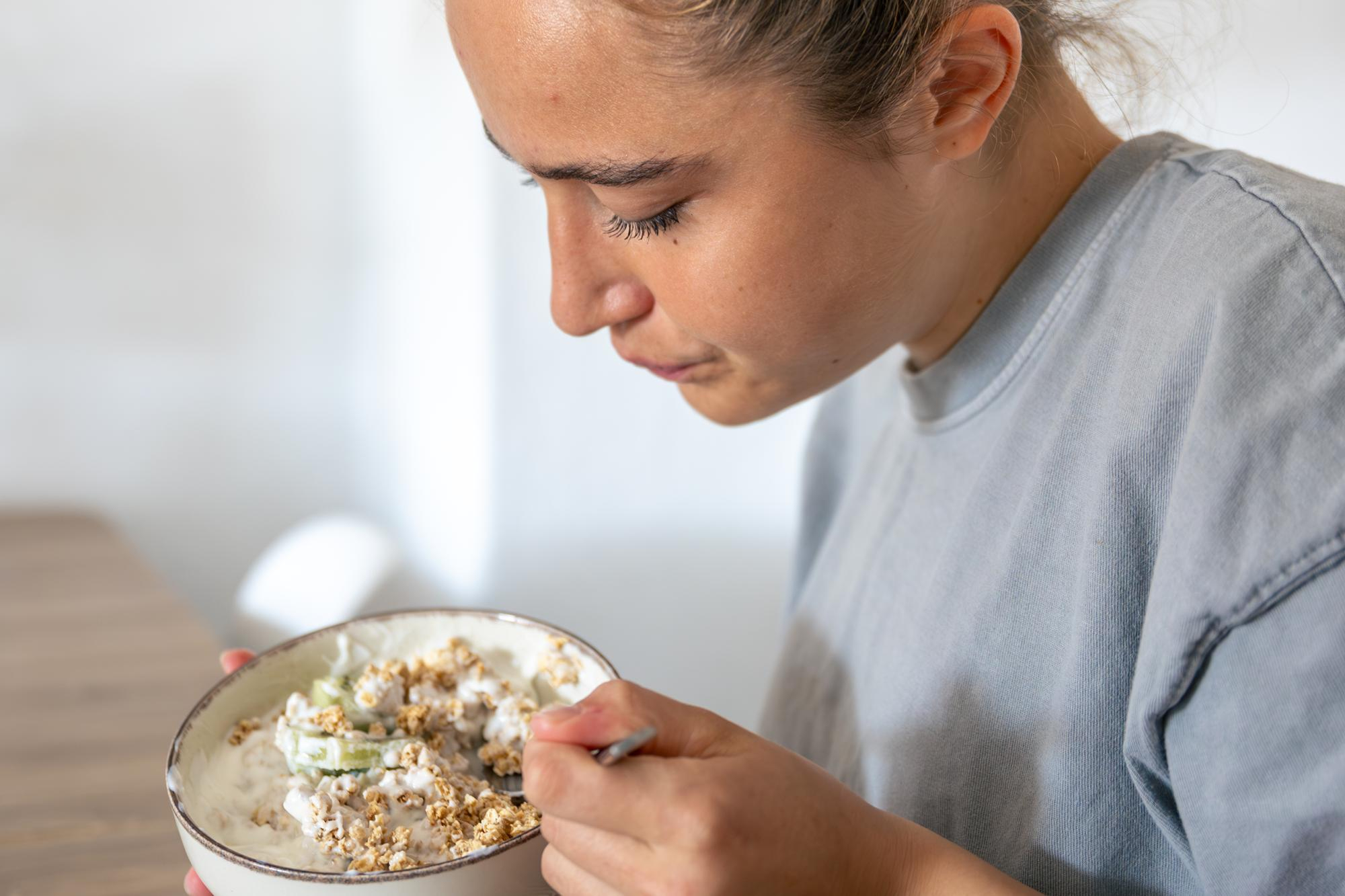Mulher jovem com cabelo preso comendo uma tigela de iogurte com oleaginosas e frutas, fontes de gorduras boas.