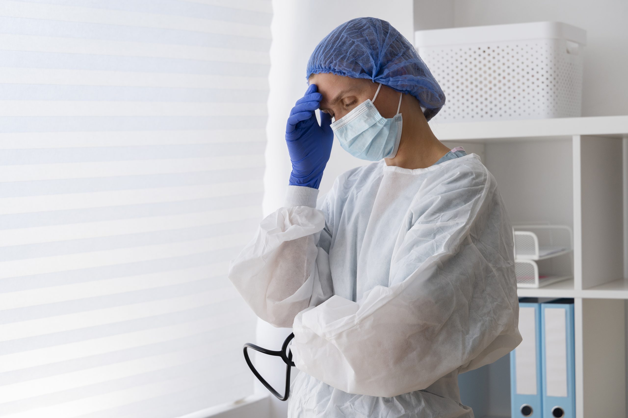 Profissional de saúde usando máscara, touca e EPI com expressão de cansaço, representando risco de alergia ocupacional em ambiente hospitalar.