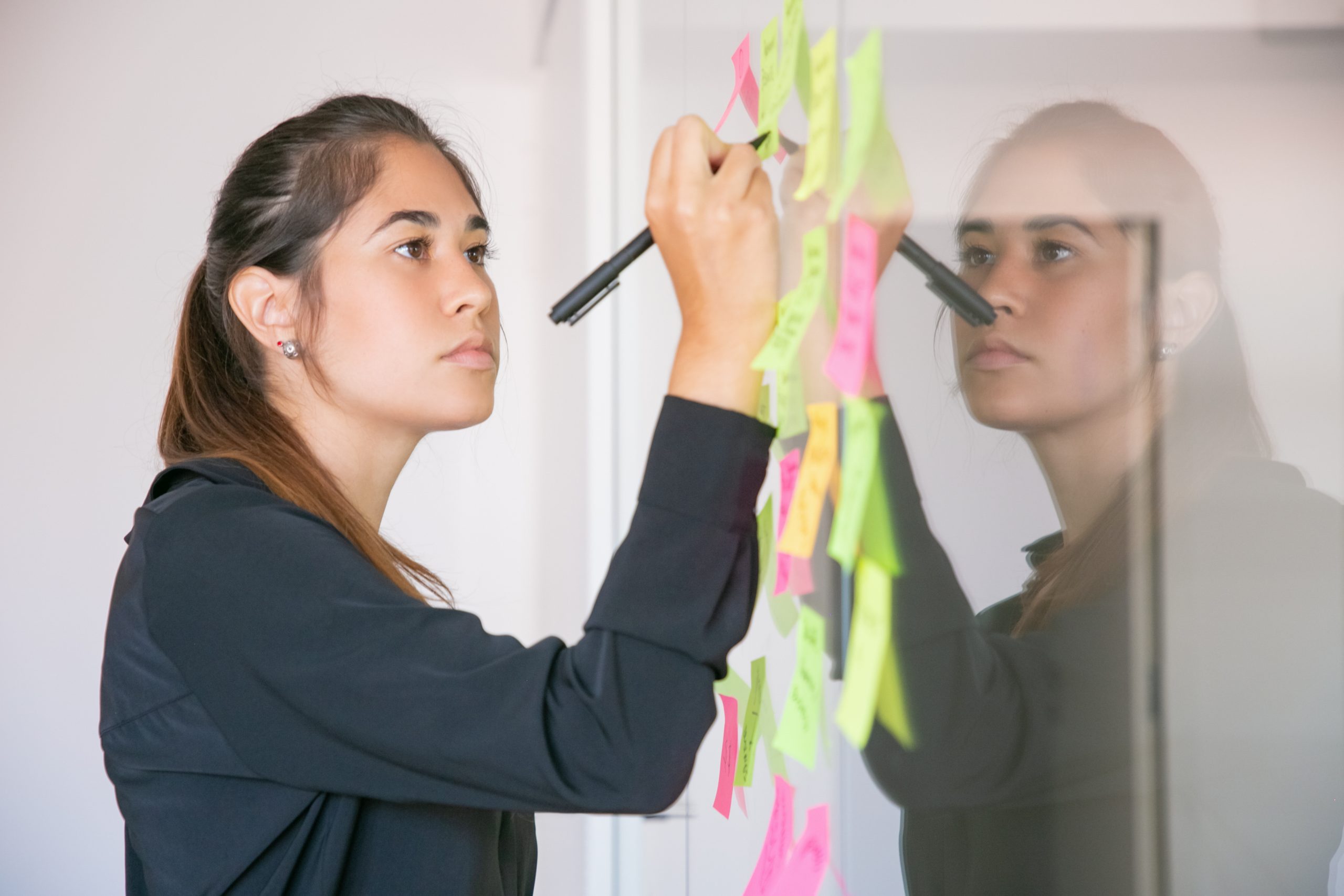 Mulher escrevendo em post-its presos a um quadro de vidro, representando o perfeccionismo, a organização excessiva e a busca constante por resultados impecáveis no ambiente de trabalho.