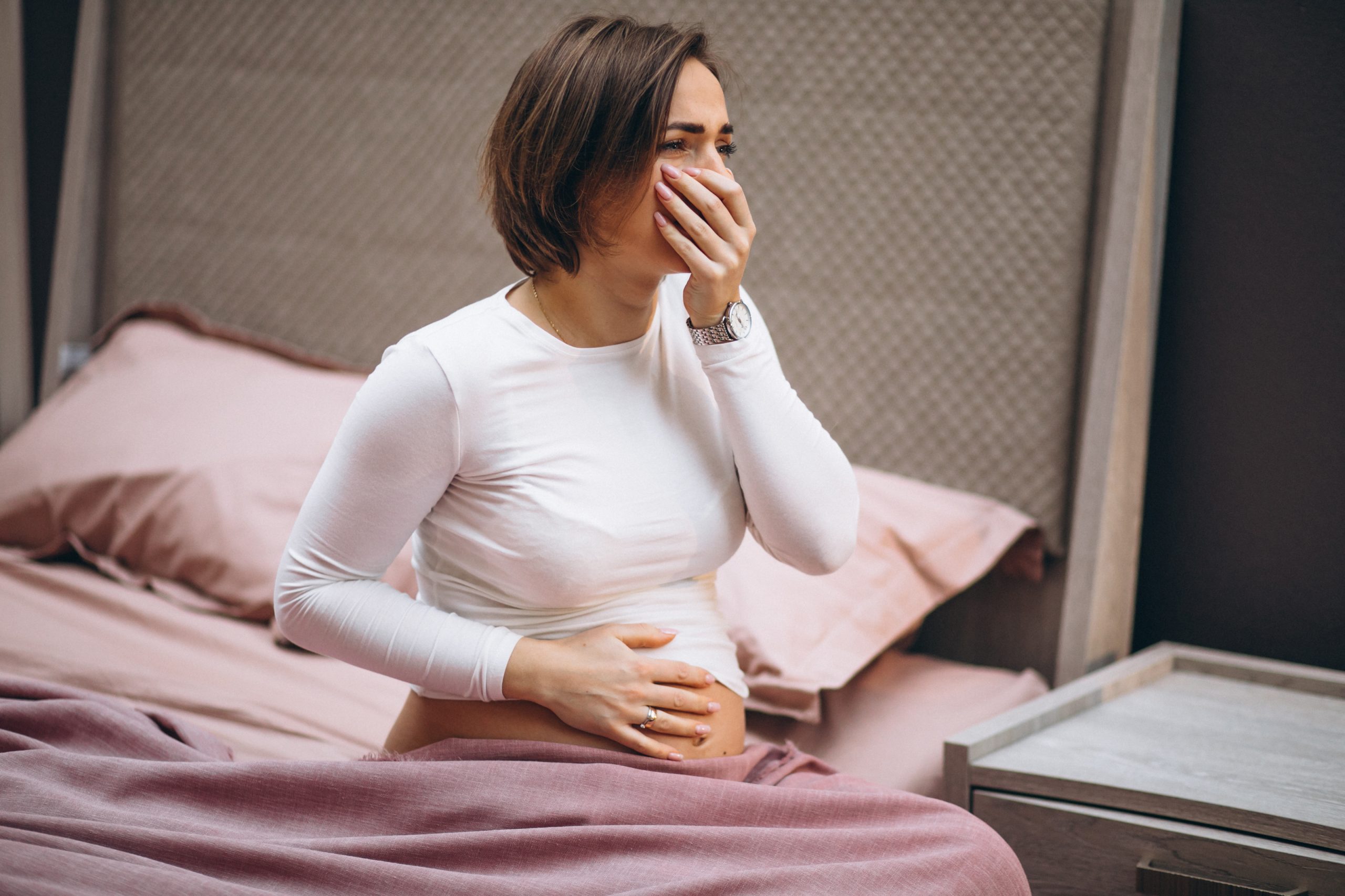 Gestante sentada na cama com a mão na barriga e apresentando mal-estar durante a gravidez, possível sintoma de toxoplasmose.