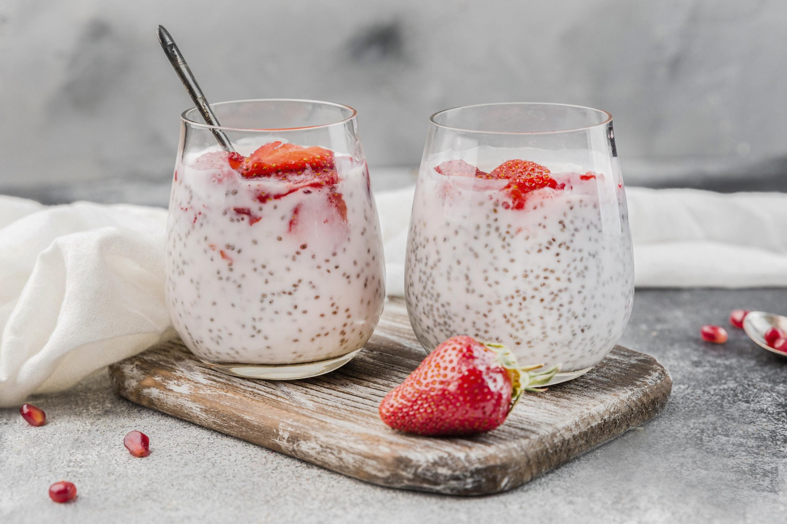 Dois copos de iogurte com sementes de chia e pedaços de morango, acompanhados de um morango fresco sobre uma tábua de madeira.