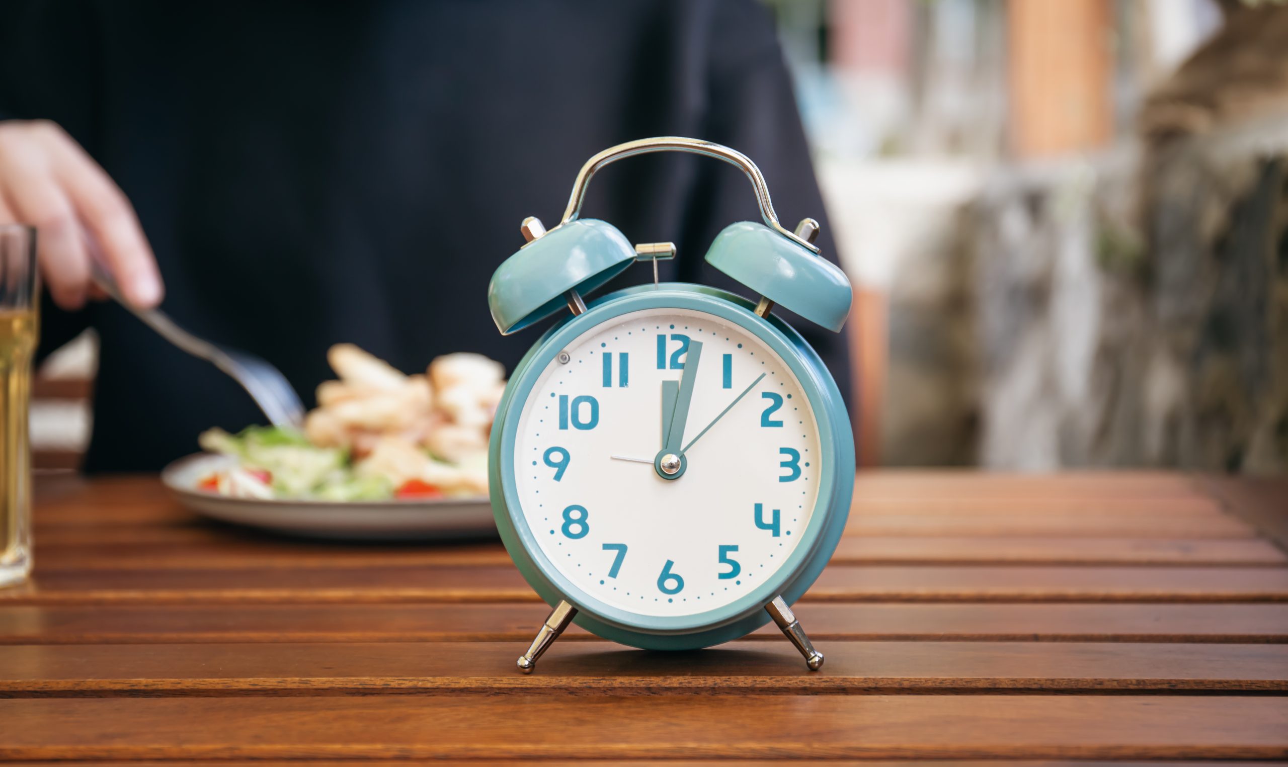 Relógio despertador sobre uma mesa, com uma pessoa fazendo uma refeição ao fundo, representando os períodos definidos para comer e ficar sem se alimentar durante o jejum intermitente.