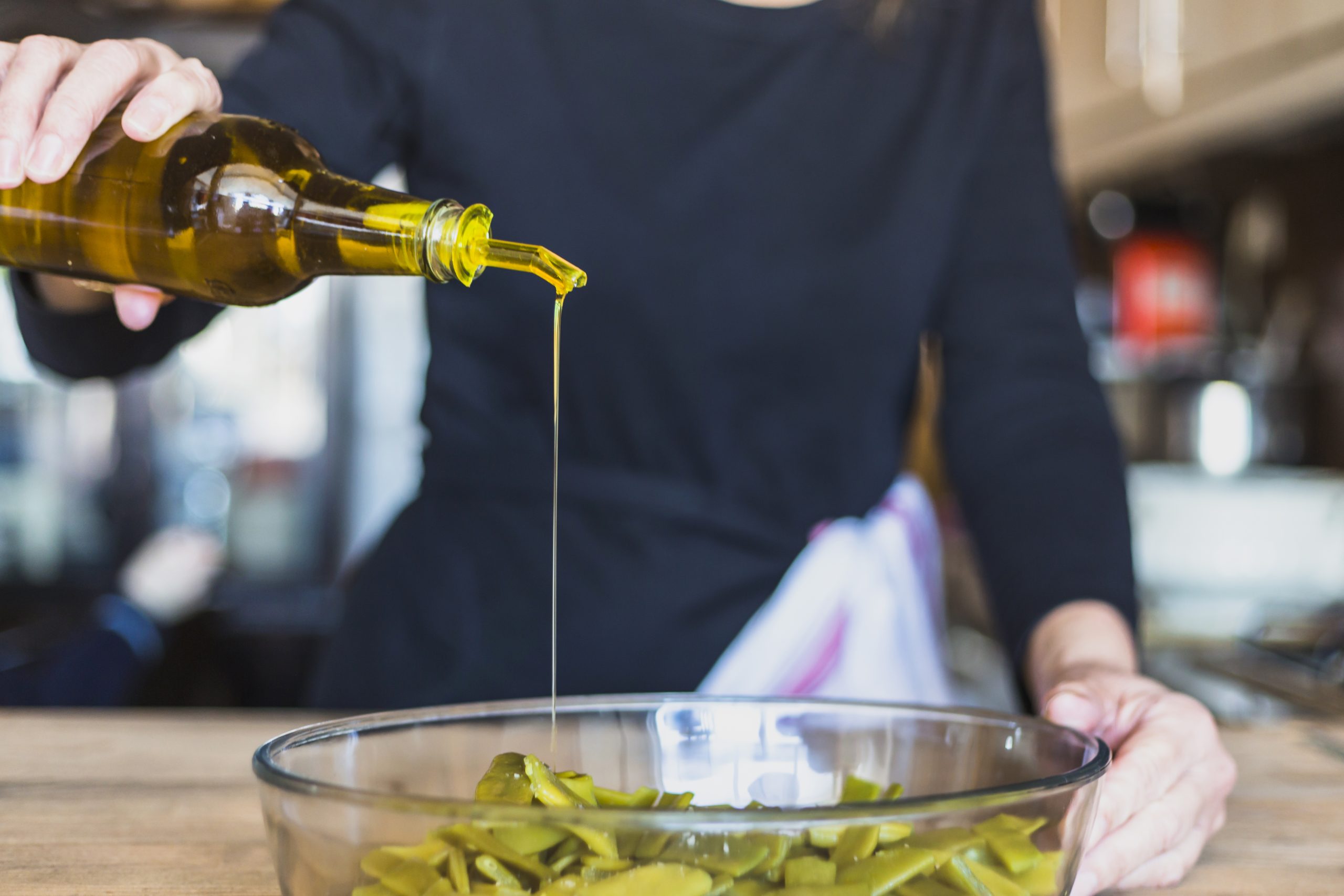 Pessoa despeja azeite de oliva extravirgem de uma garrafa sobre uma salada com vegetais verdes em uma tigela de vidro, durante o preparo de uma refeição na cozinha.
