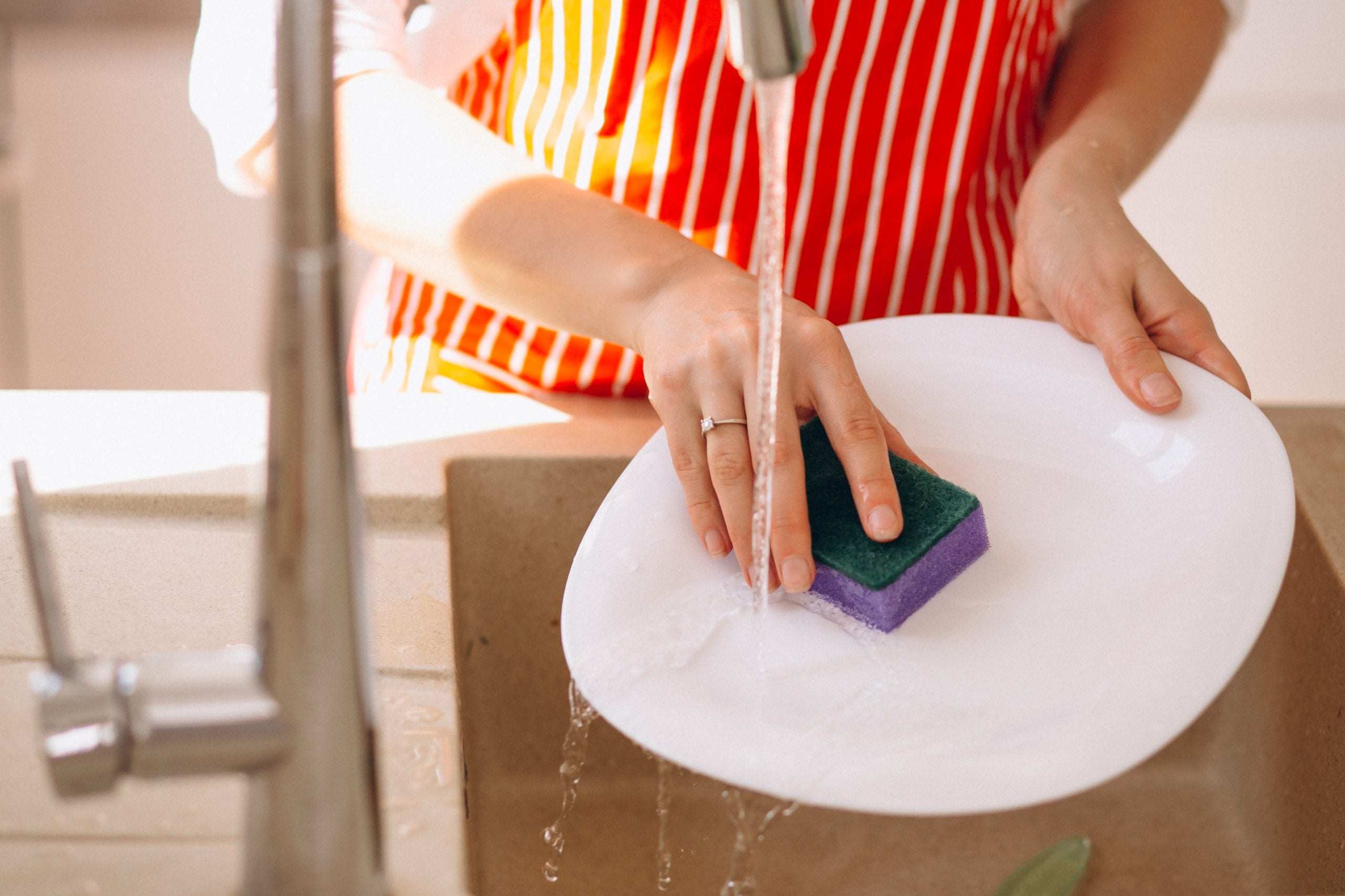 Pessoa lava um prato branco com uma esponja de cozinha verde e roxa sob a água corrente da torneira.