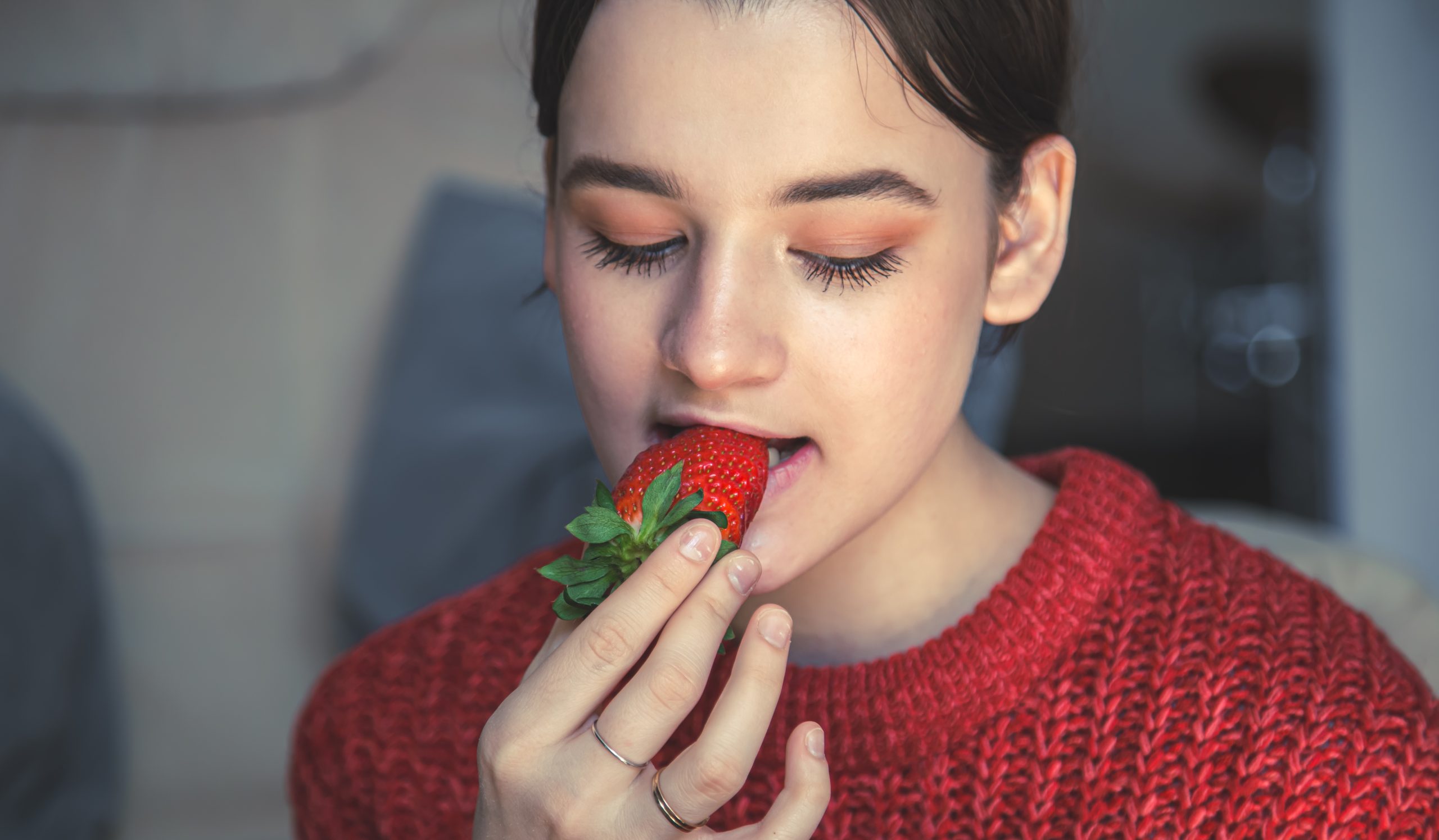Mulher segurando um morango vermelho próximo à boca, prestes a comer a fruta, que é fonte de vitamina C, fibras e antioxidantes.