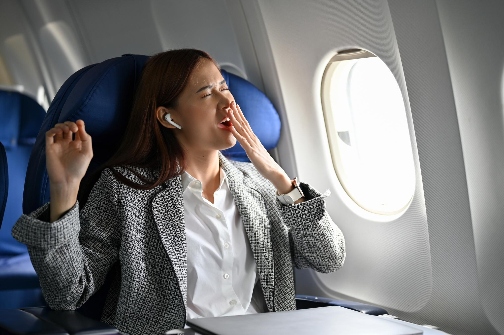 Passageira bocejando em avião enquanto ajusta o relógio para um novo fuso horário, representando o jet lag.