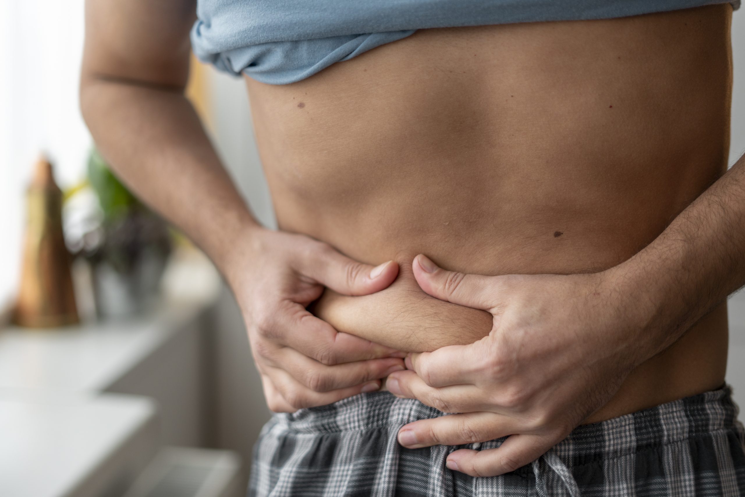 Pessoa segurando a parte inferior da barriga com as duas mãos, mostrando leve acúmulo de gordura abdominal. A imagem ilustra o estômago alto, o inchaço na região abdominal e as possíveis causas do aumento do volume da barriga.