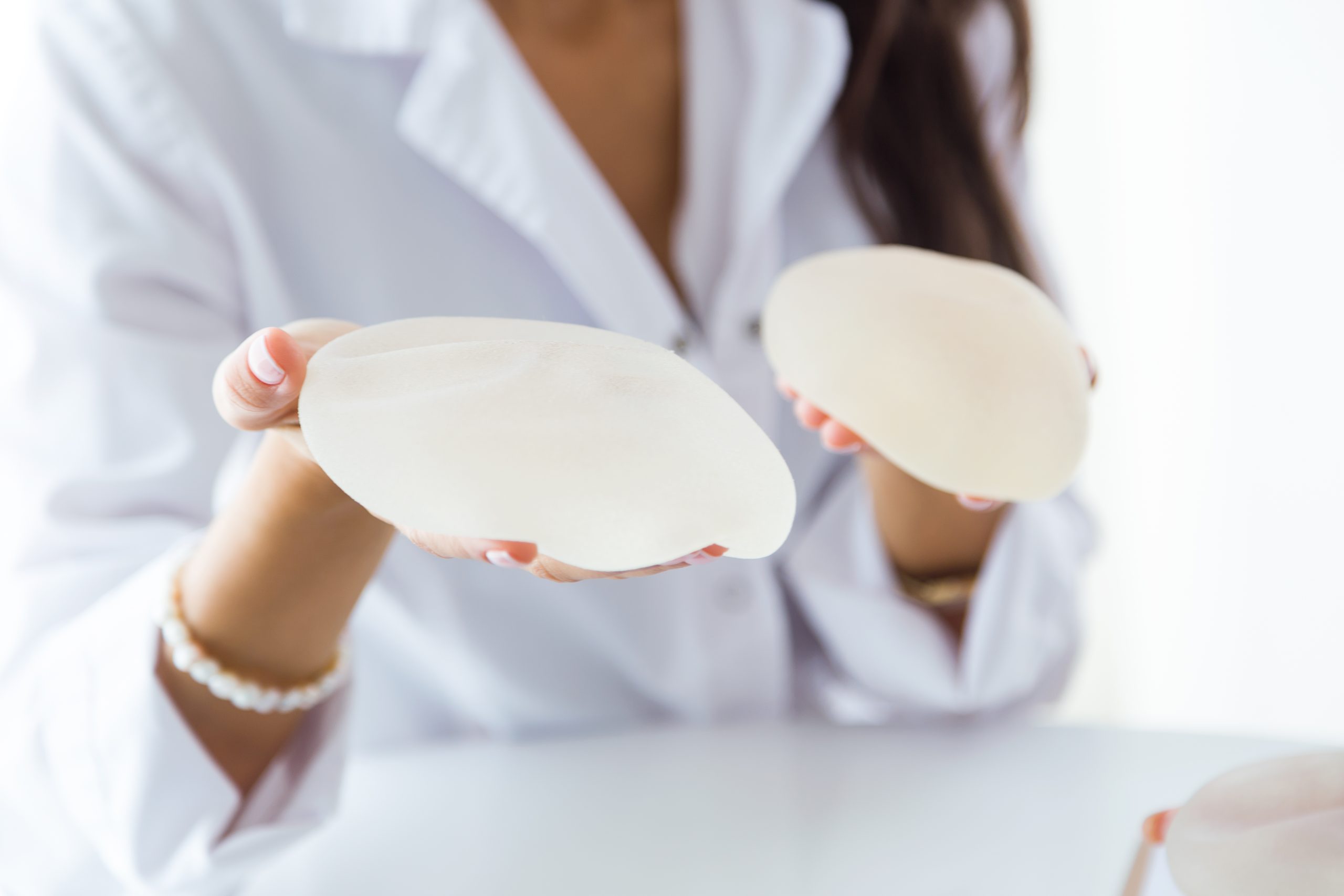 Profissional de saúde de jaleco branco segura duas próteses de silicone mamárias, uma em cada mão, durante atendimento em consultório.